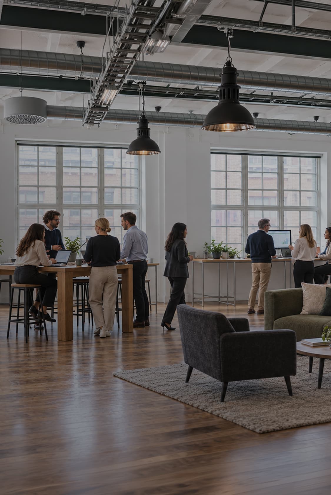 Colleagues talking and working at tall tables in a bright open-plan office with exposed ceiling pipes and a sofa