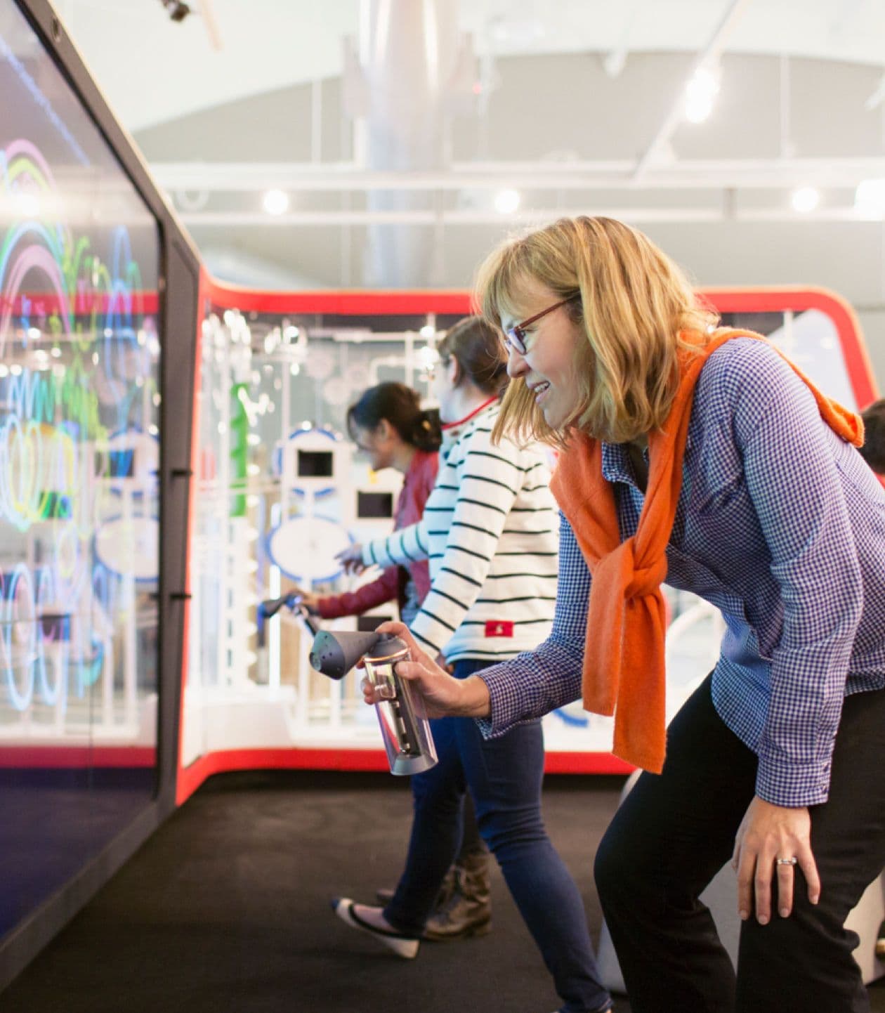 Smiling woman in glasses and orange scarf interacts with a glowing, colorful exhibit using a spray-like device.