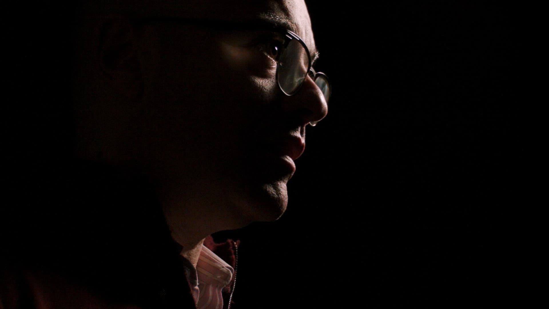 A man's face in profile, wearing glasses, half-lit against a dark background, casting deep shadows.