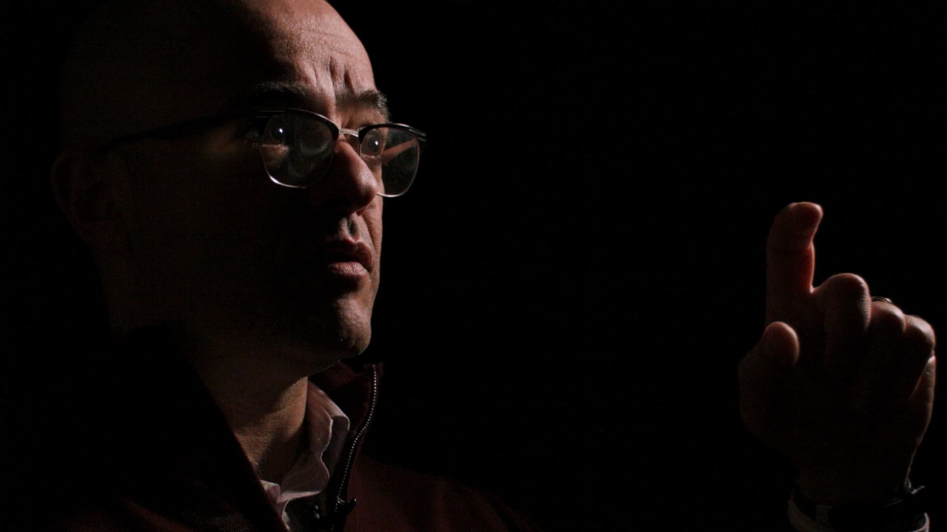 A man with glasses, dramatically lit from the side, points his index finger upwards against a black background.