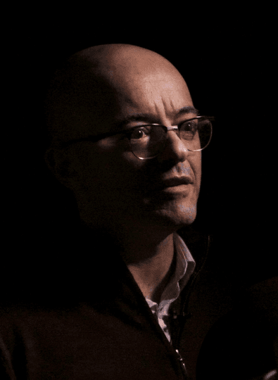 Dark portrait of a bald man in glasses gesturing with one hand, lit softly against a black background