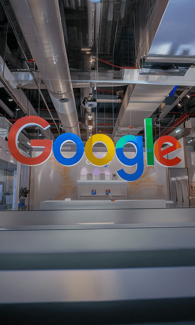 Large, colorful Google logo hanging in a modern office or retail space with exposed ceiling ducts and a white counter.