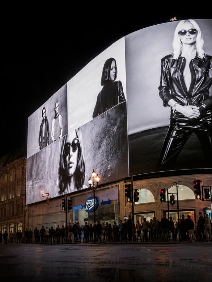 Digital billboards showing black-and-white fashion campaign imagery above crowds at Piccadilly Circus, London, at night