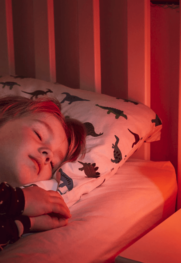A young child with closed eyes sleeps soundly in a crib, bathed in soft red light. A dinosaur-patterned pillow supports their head.