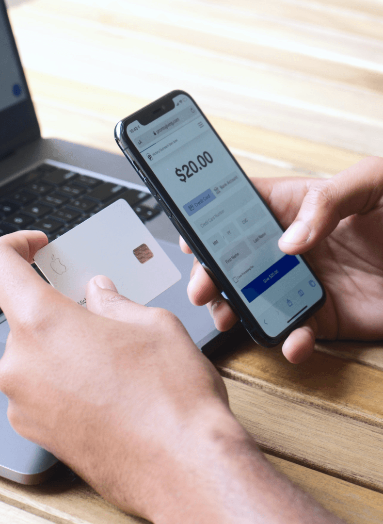 Hands holding a phone showing a $20.00 card payment form and a white bank card, beside a laptop