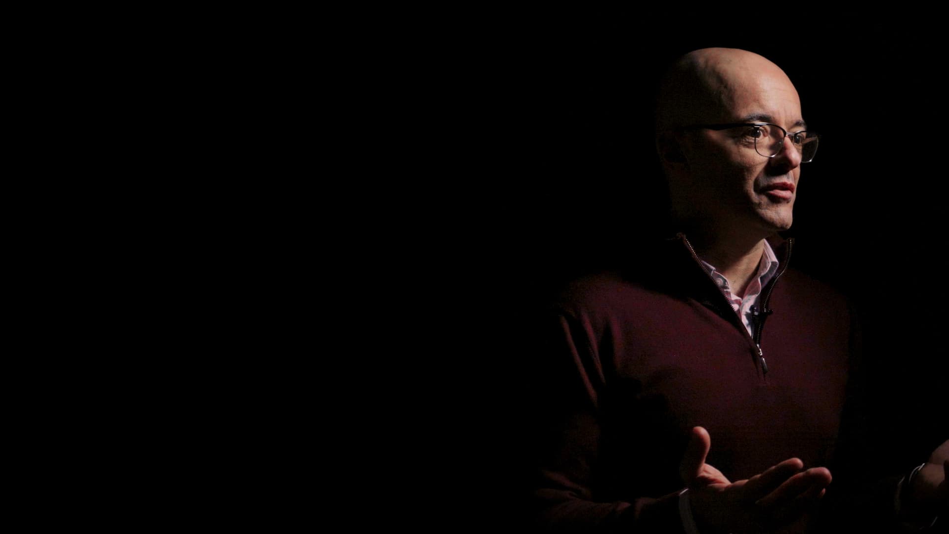 Gilad G. Amir in glasses and a red sweater, gesturing as he emerges from darkness, looking to the right