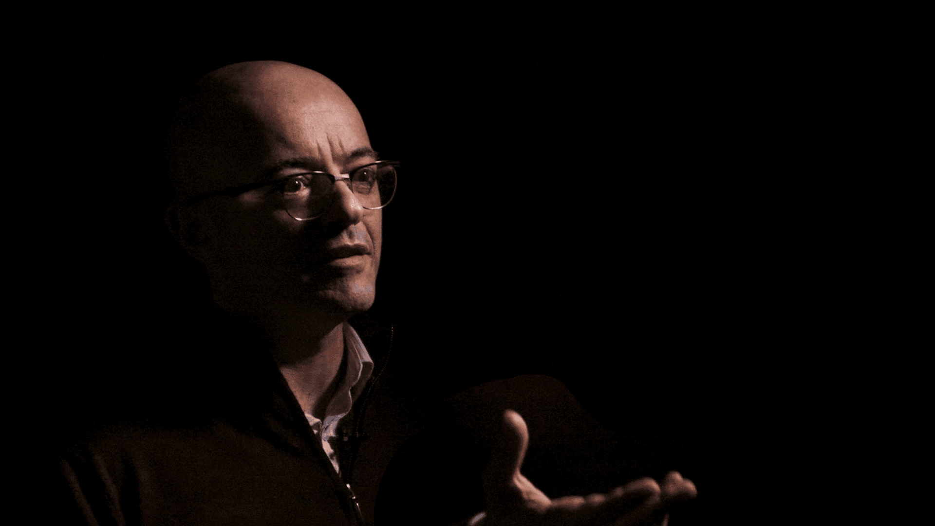 A person with glasses gestures with one hand while partially lit, standing against a dark background.