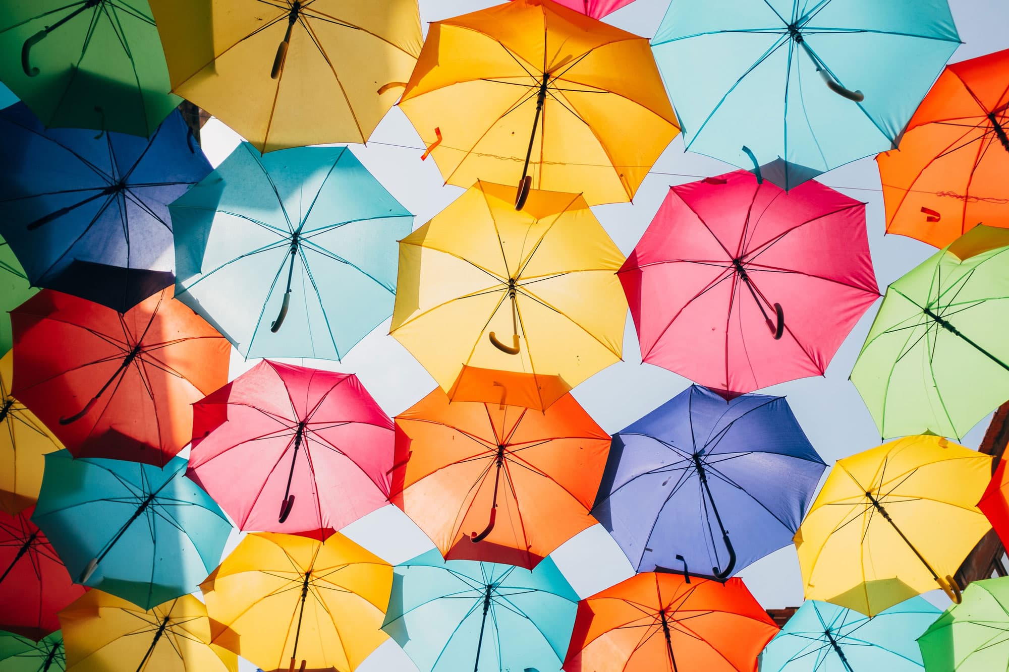 A vibrant canopy of numerous colorful open umbrellas in shades of yellow, orange, pink, blue, green, and red, viewed from below.