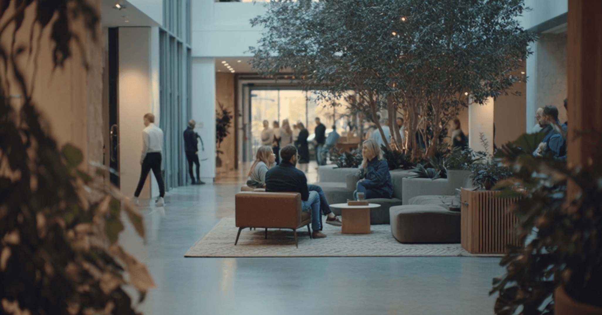 People sitting and conversing in a modern, indoor atrium with large plants, natural light, and a sleek design. Others walk by in the background.