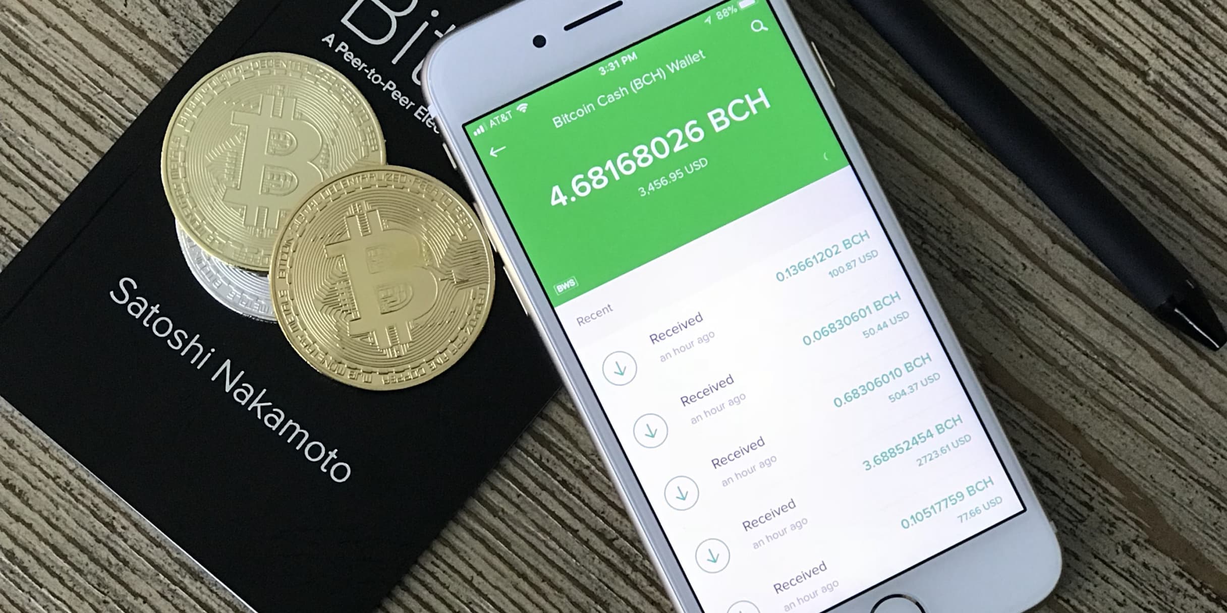 Smartphone showing a Bitcoin Cash wallet balance beside two gold coins on a book by Satoshi Nakamoto