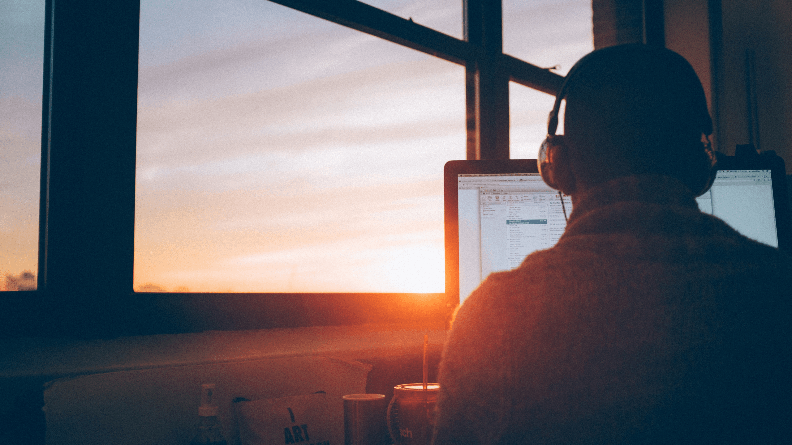 Person with headphones working on a computer, silhouetted against a vibrant sunset visible through a large window.
