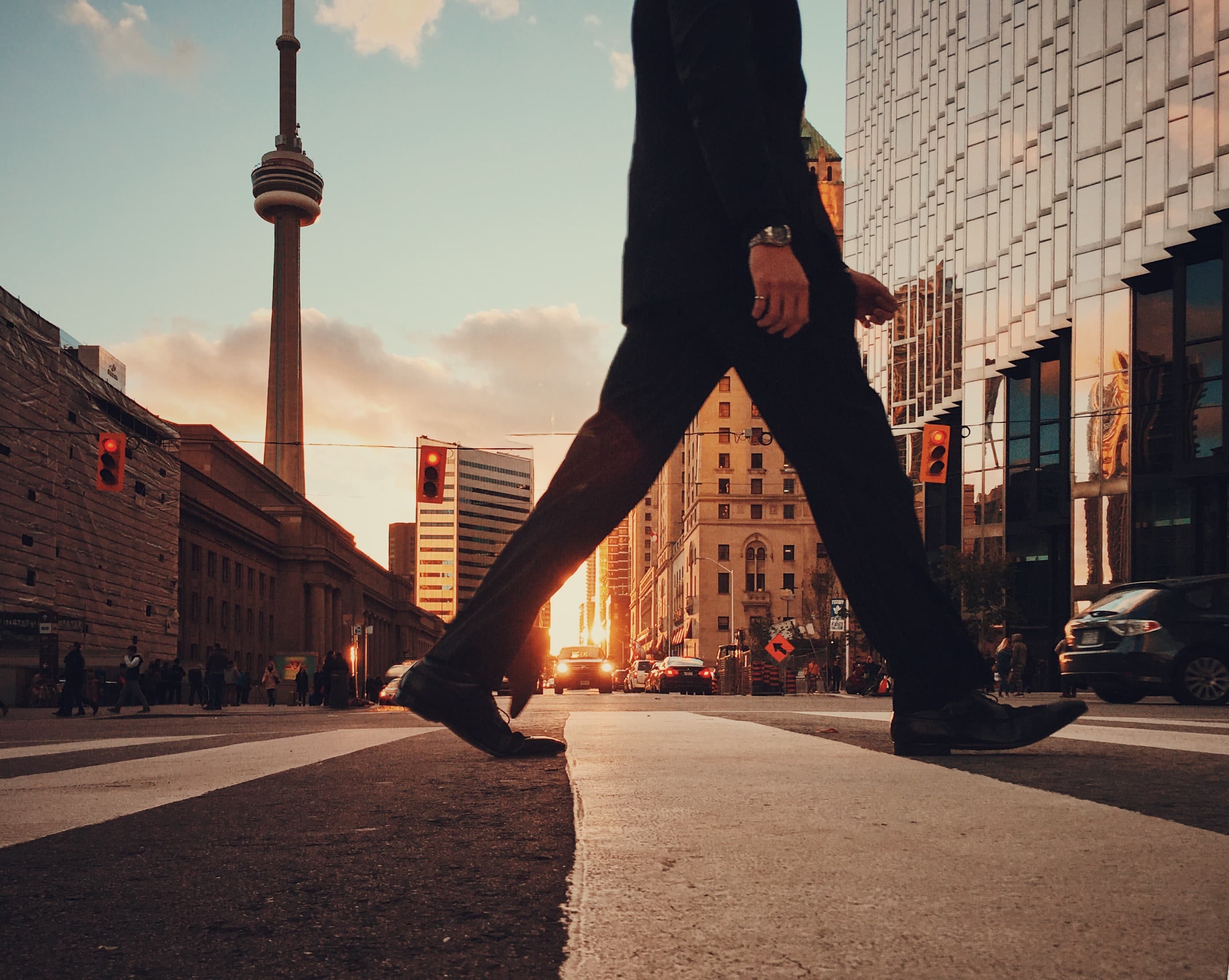 Person in dark trousers walking across a zebra crossing in a city street at sunset, with a tall tower behind