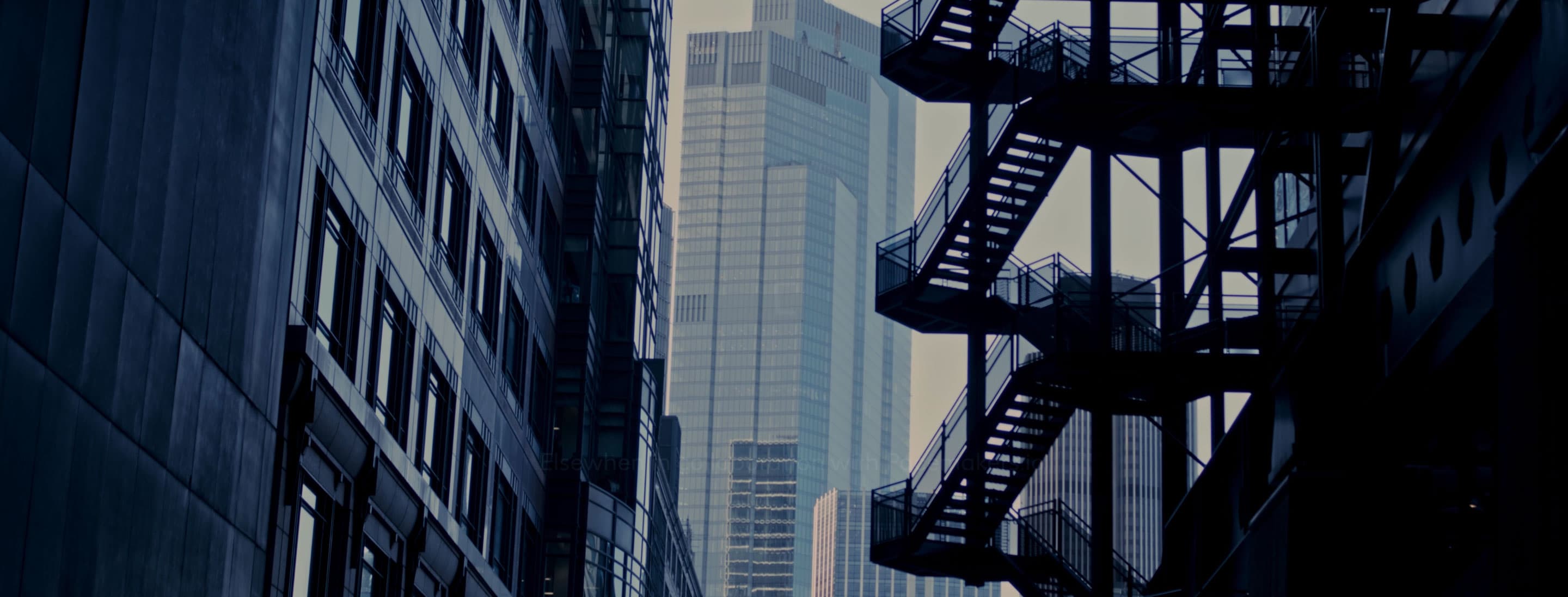 Looking up a dark, narrow city alley between a building and a fire escape, with a tall, modern skyscraper in the background.