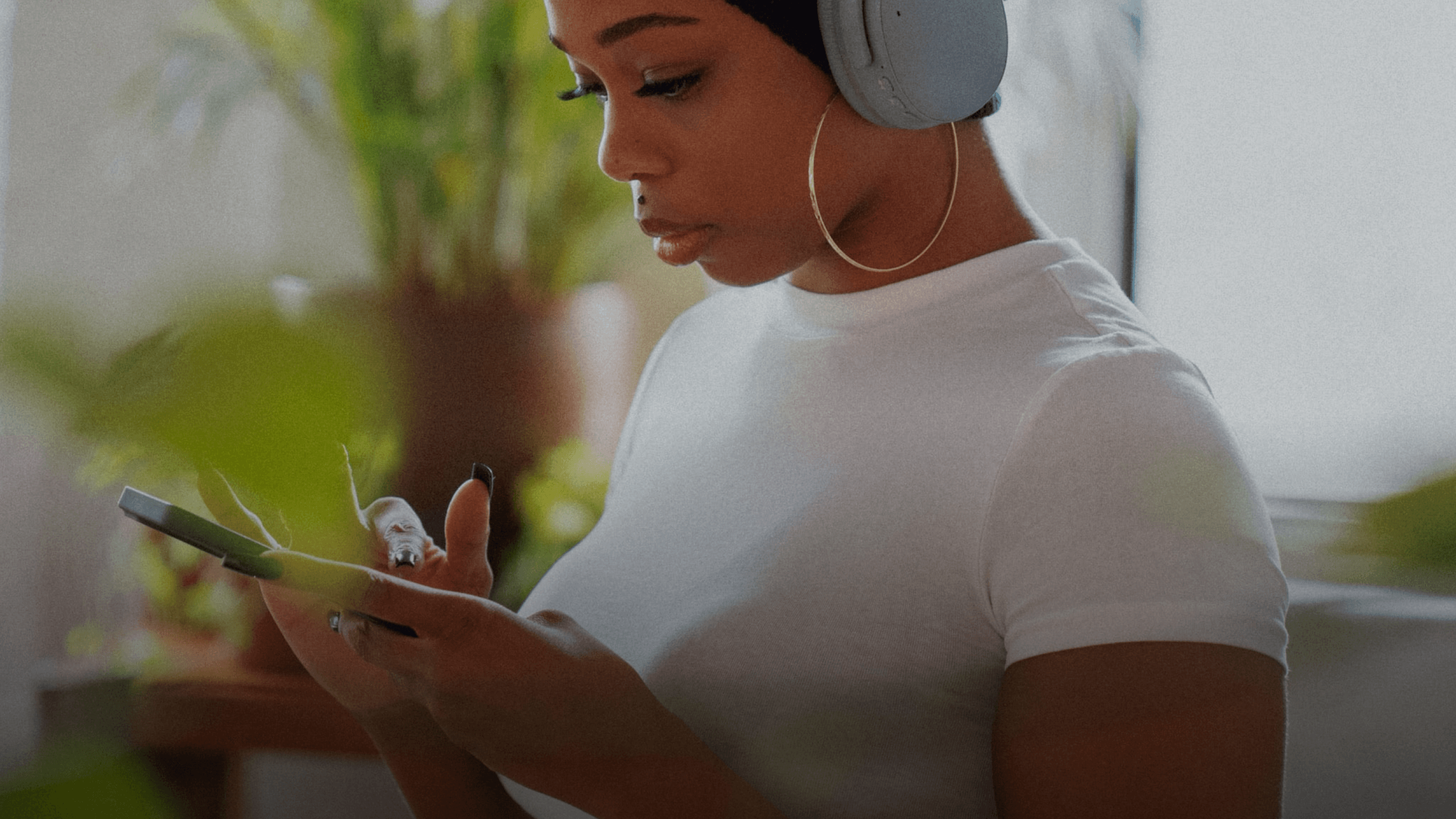 A young Black woman wearing grey headphones and a white t-shirt, looking at her smartphone. Green plants are blurred in the background.