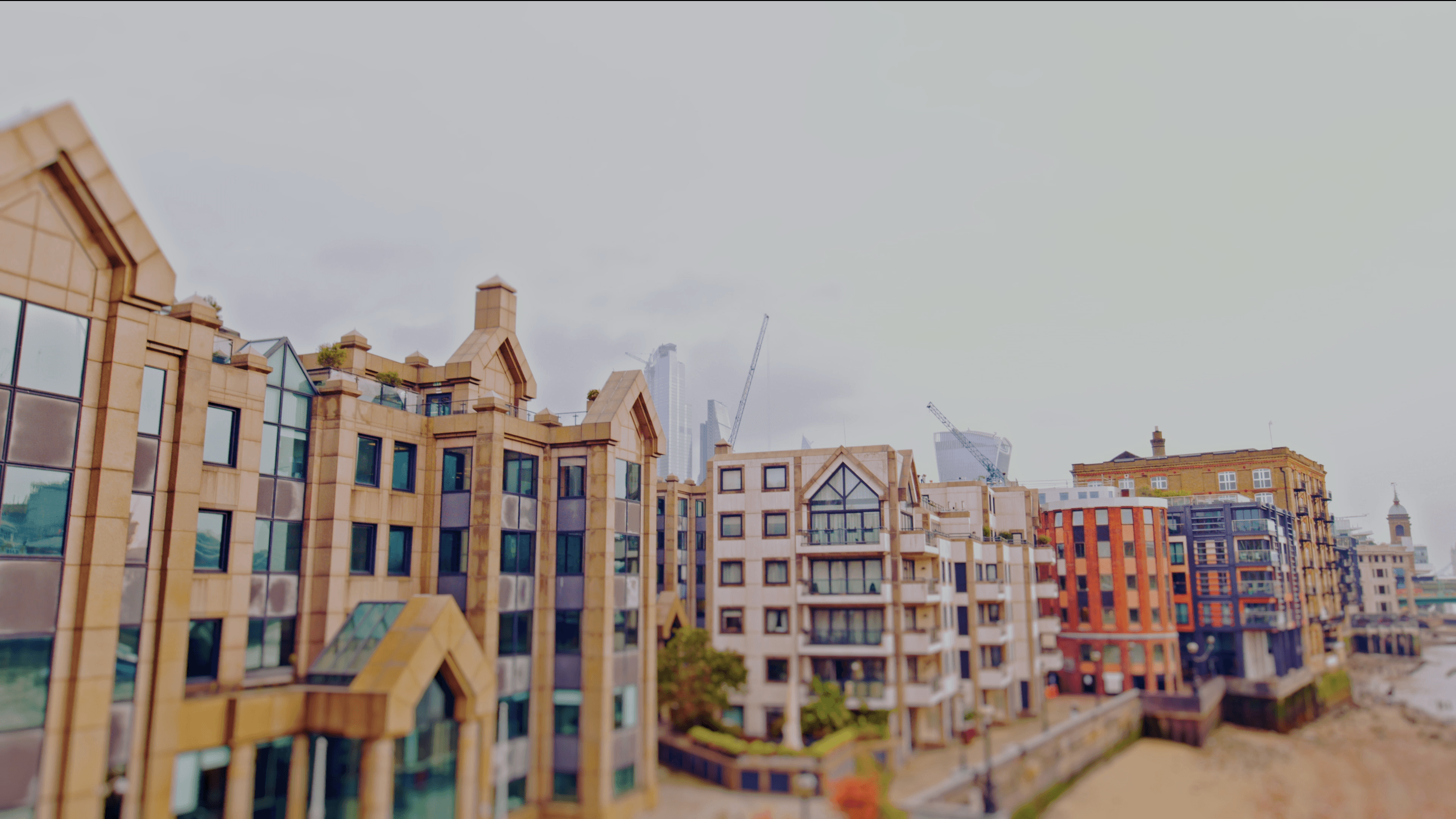 Tilt-shift photo of diverse buildings lining a city riverbank, from tan to colorful, with distant cranes under an overcast sky.