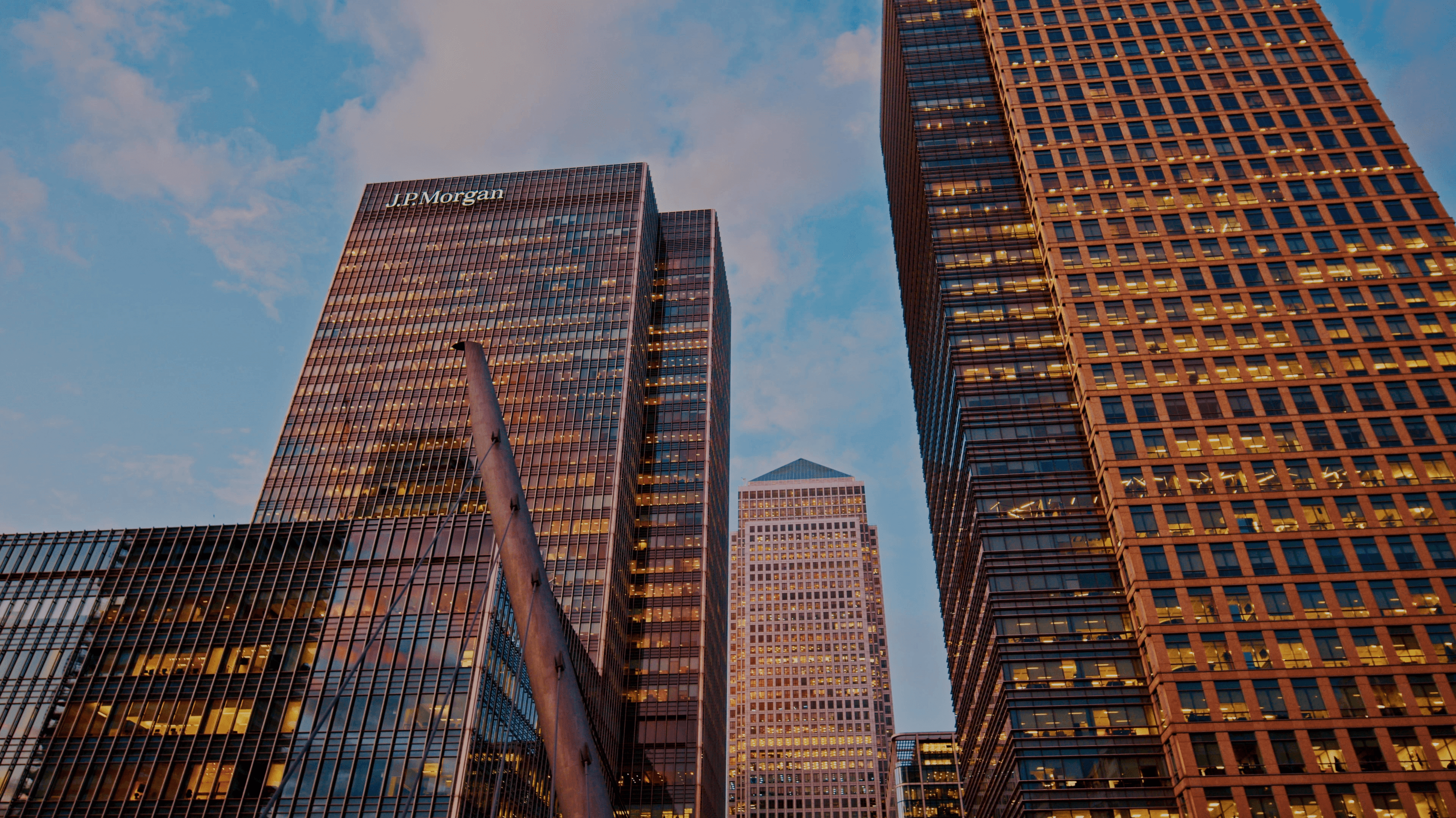 Modern skyscrapers, including the J.P. Morgan building with illuminated windows, at dusk against a cloudy blue sky.