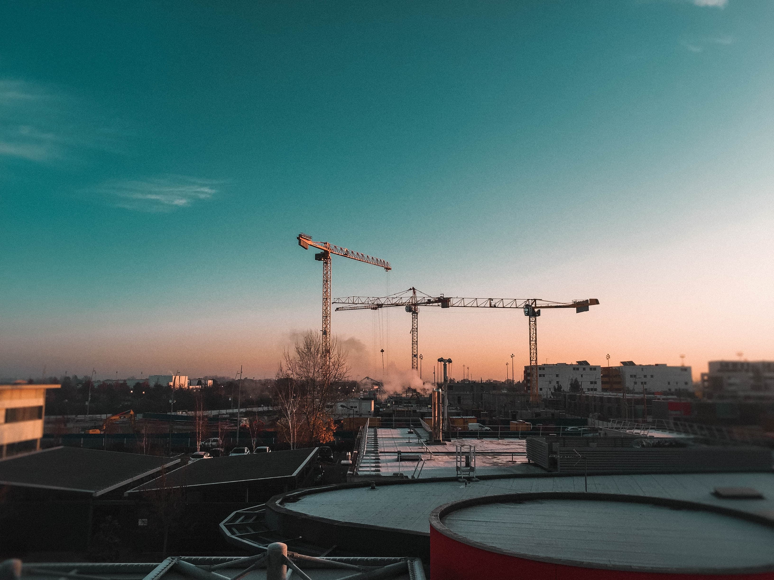 Construction cranes on a building site at sunset under a teal sky, with rooftops in the foreground