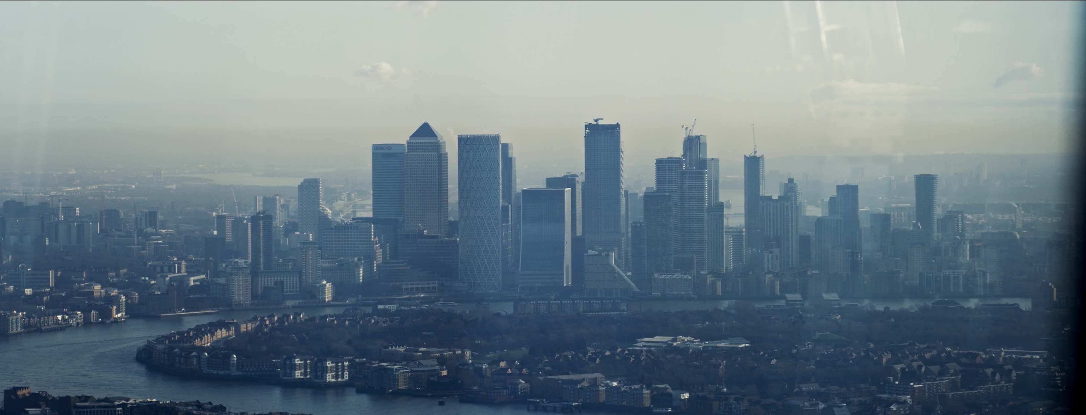 Aerial view of London's Canary Wharf skyline, with modern skyscrapers and the River Thames winding through a hazy landscape.