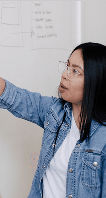 A woman in clear glasses and a denim jacket points towards a whiteboard with writing, looking engaged in discussion.