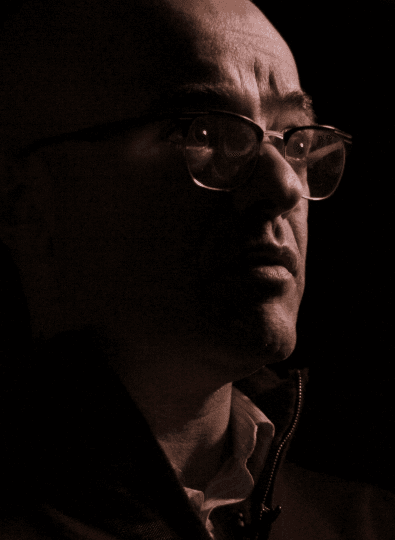 Close-up profile of a man with glasses, lit dramatically from the side against a dark background, with a furrowed brow.