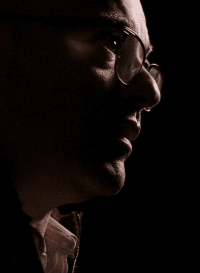 Dark side-profile portrait of a bald man in round glasses, lit softly against a black background