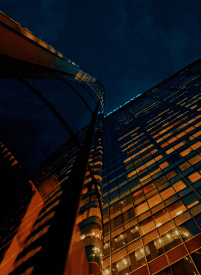 Tall glass skyscrapers with lit orange windows seen from below against a dark blue night sky