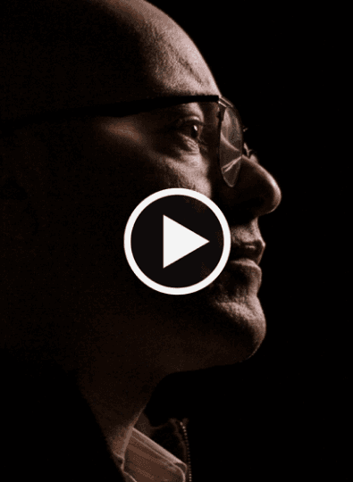 Side profile of a bald man with glasses in dark lighting, with a play button over the image