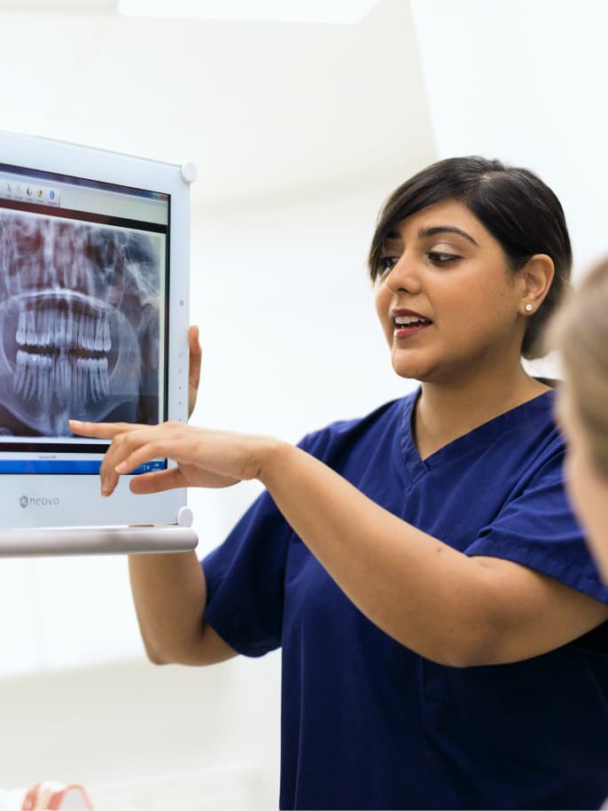 A dentist in blue scrubs explains a dental X-ray to a patient, pointing to details on a computer screen.