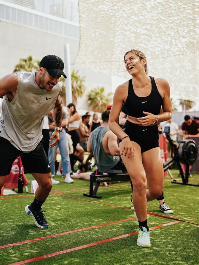 Happy man and woman in athletic wear exercising outdoors on green turf, with others in the background.