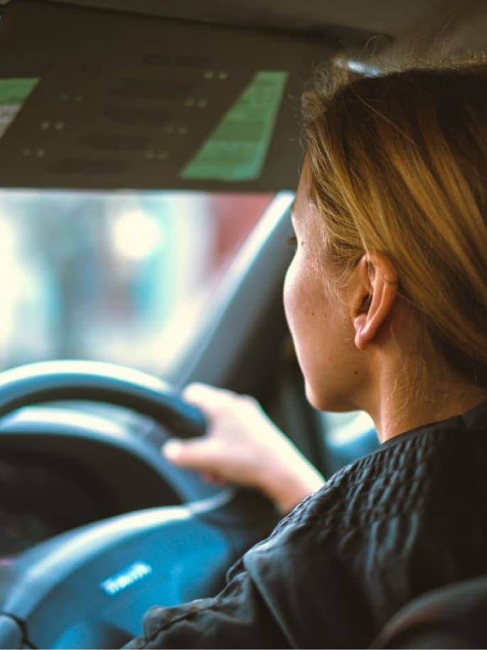 Blonde woman driving a car, viewed from behind her right shoulder. Her hand is on the steering wheel, with blurred city lights outside.