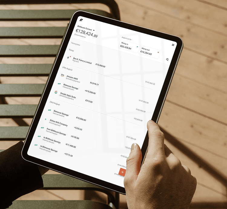 A person holding a tablet displaying a financial summary with transaction details and balances in euros, alongside a wood floor background.