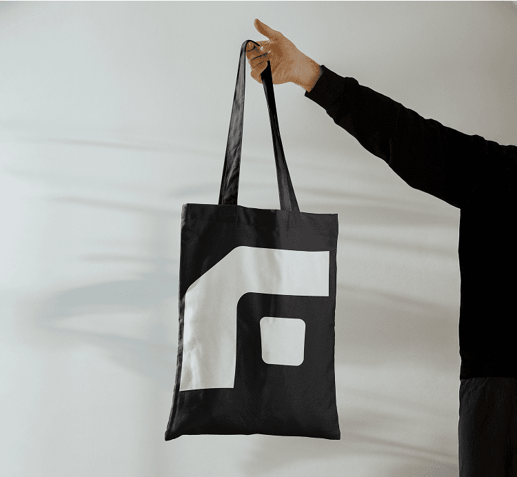 A hand holding a black tote bag with a bold, abstract white design on it, set against a plain light background.