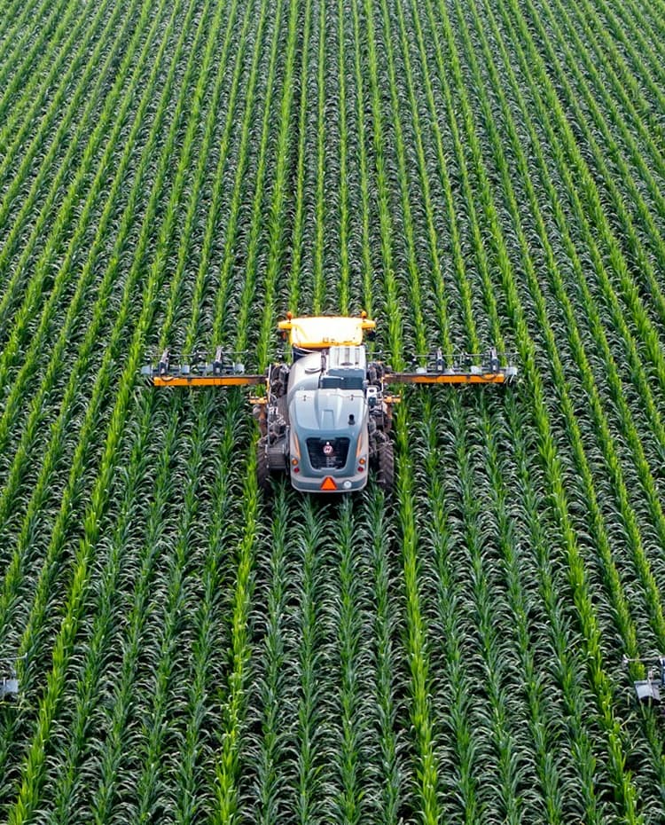 Aerial view of a tractor spraying crops in a green field, with precisely aligned rows stretching into the distance.