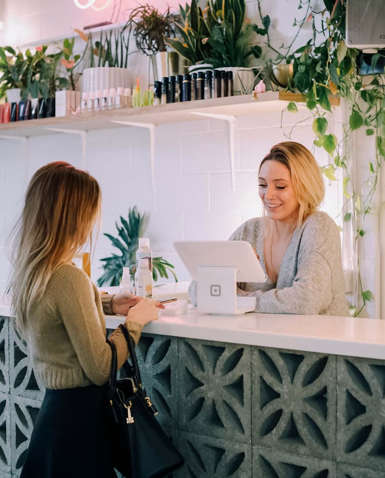 Customer paying a smiling shop assistant at a counter with a tablet till, with plants and shelves of products behind