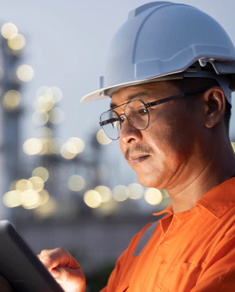 An engineer in a hard hat and orange hi-vis using a tablet at an industrial facility at dusk.