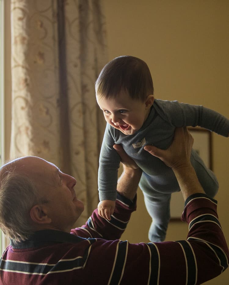 A joyful older man holds a giggling baby high in the air, looking up at the baby who smiles back.