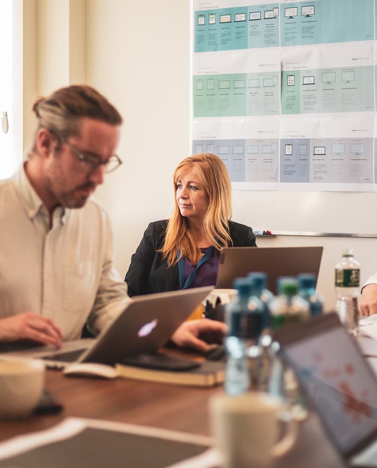 People in a meeting room working on laptops, with business charts on the wall behind them.