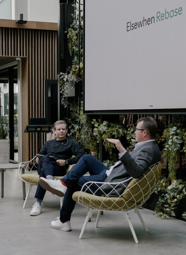 Two men in conversation seated in chairs at an Elsewhen Rebase event, in front of a screen reading "Elsewhen Rebase" and a wall of plants