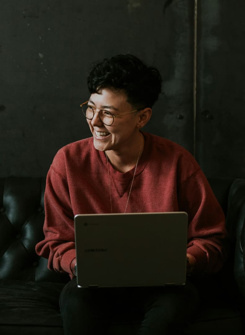 A person with short, curly hair and glasses, wearing a red sweatshirt, laughs while holding a Samsung laptop on a dark couch.