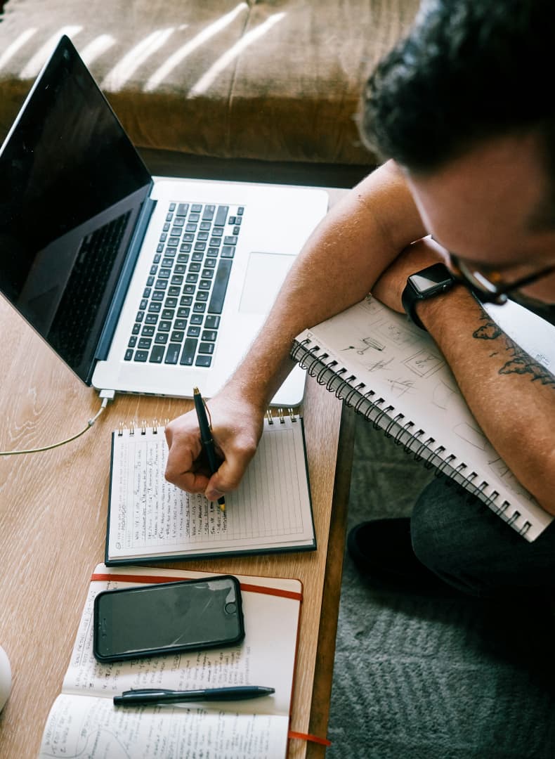 Overhead view of a man writing in a notebook with a pen. A laptop, smartphone, and other notebooks are on a wooden table.