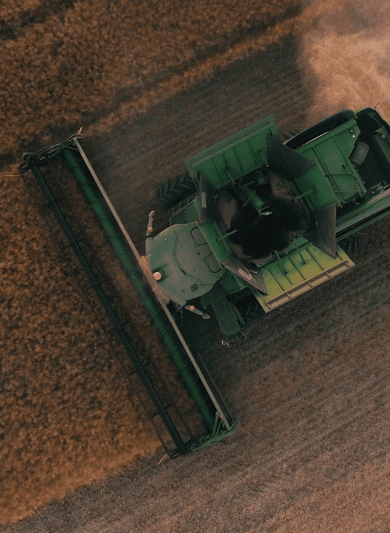 Aerial view of a green combine harvester cutting a golden crop field, with dust behind it