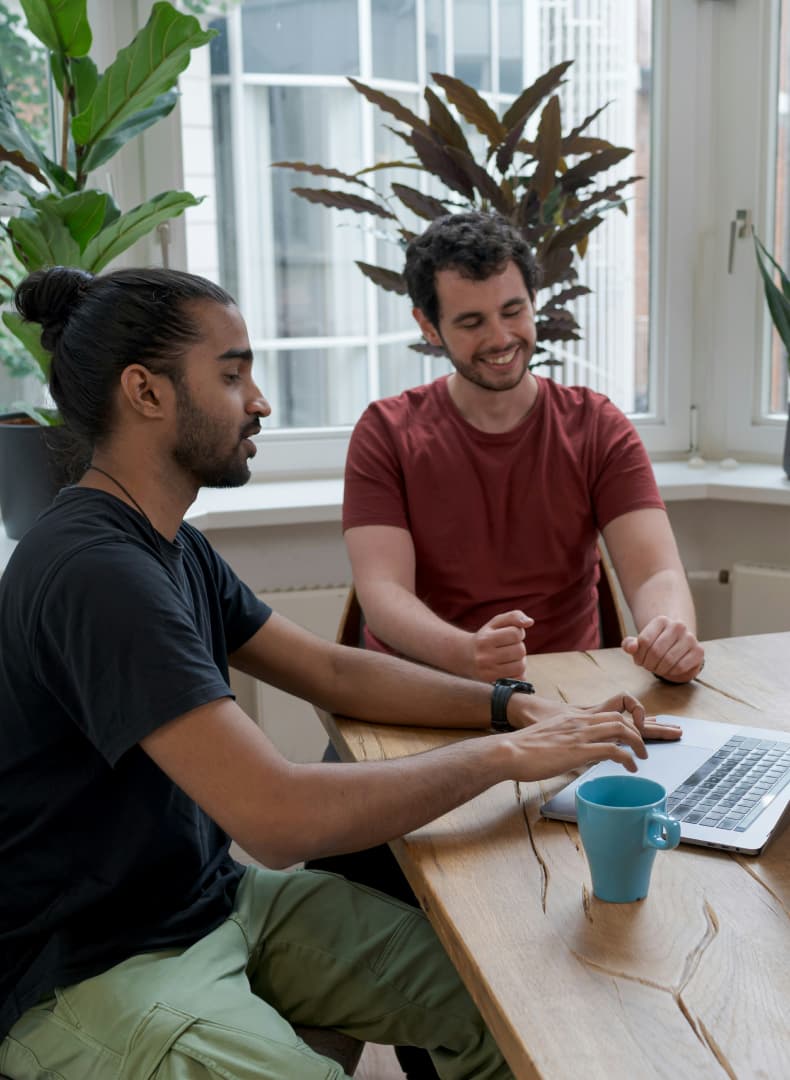 Two men at a wooden table. The man with a bun uses a laptop, talking to a smiling man in red. Plants are in the background.