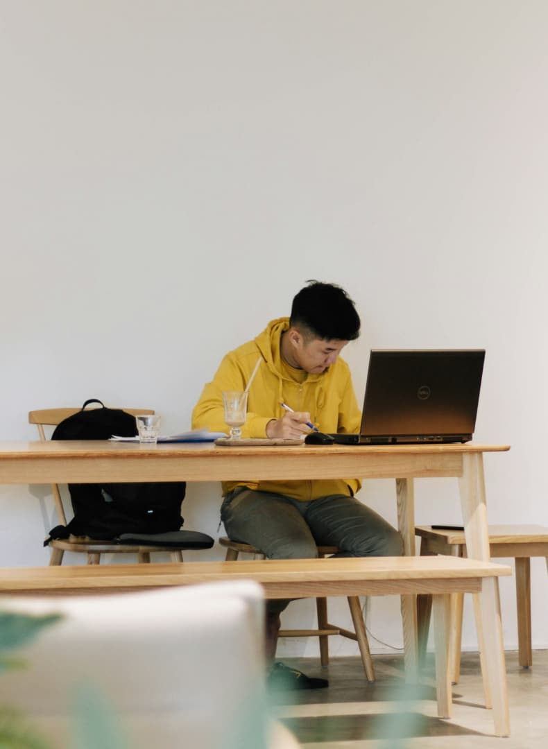 A person in a yellow hoodie works on a laptop at a wooden table, surrounded by notebooks and a glass of water.