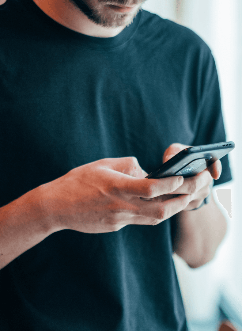 A person with a beard and dark t-shirt holds and looks at a black smartphone.