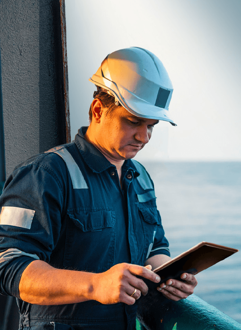 Worker in a white hard hat and navy overalls looking at a tablet on a ship deck, with the sea behind